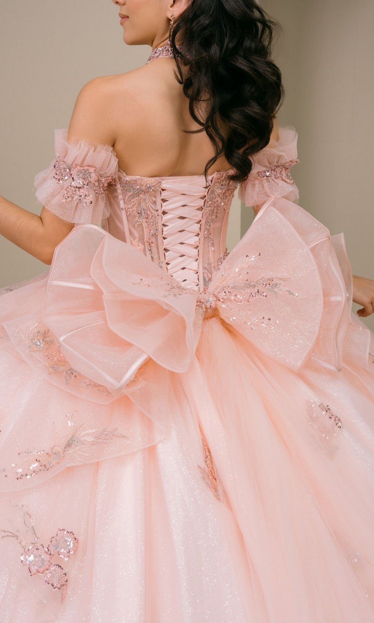 A woman wearing the Quinceanera Dress 8540JQ By Cinderella Coutureโa sparkly blush pink gown featuring off-shoulder sleeves, floral details, a corset back, and a large bowโperfect for a quinceanera ball gown look.